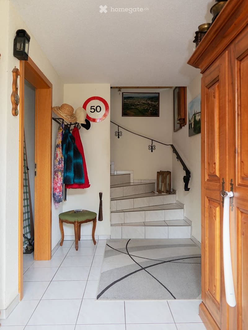 Hallway with tiled floor, wooden staircase, coat rack with hats and jackets, small green stool, and framed pictures on the wall.
