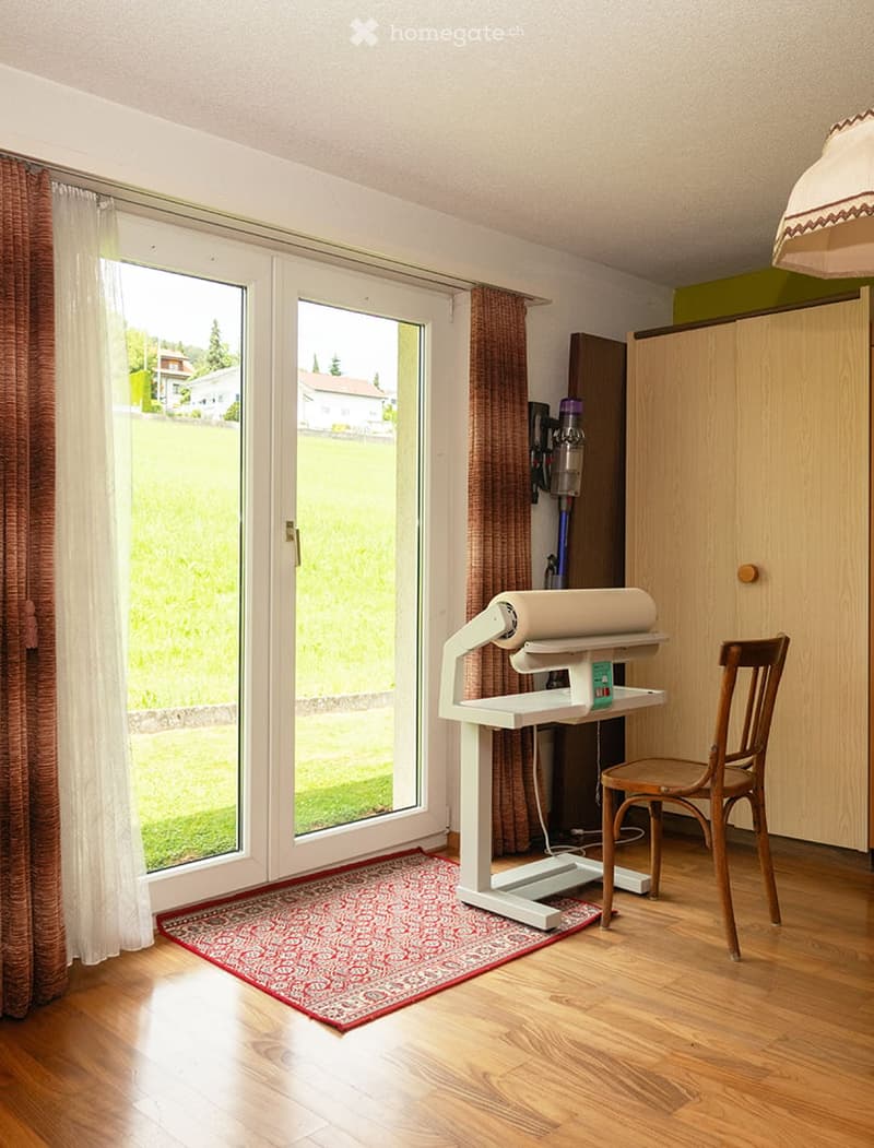 Bright room with wooden flooring, large windows offering a view of a green landscape, a sewing machine on a table, a wooden chair, a cabinet, and curtains.
