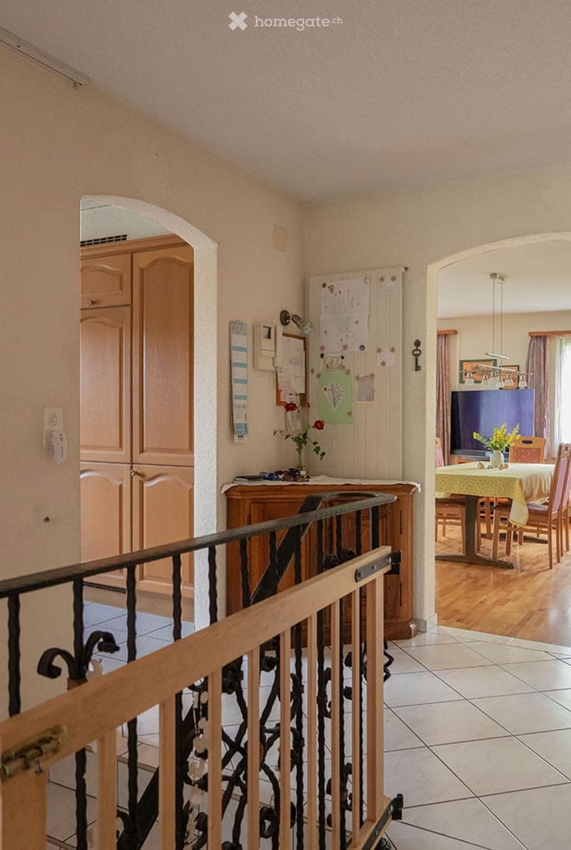 A well-lit hallway with a wooden railing leading to a dining area. The hallway features light-colored walls, a tiled floor, and a small wooden cabinet with a vase of flowers on top. The dining area visible through an archway includes a table with chairs,