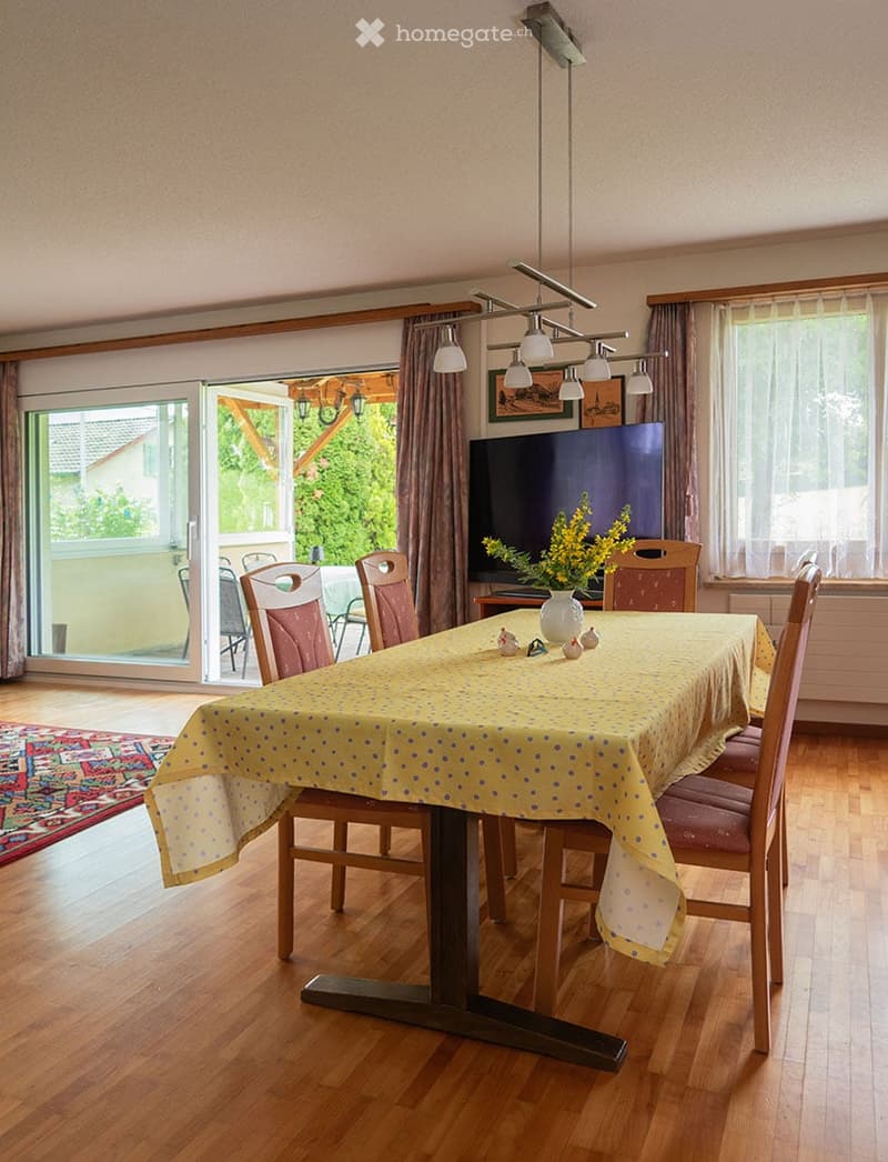 Dining room with wooden floor, large sliding glass door leading to a balcony, dining table with chairs, television, and flower vase on the table.