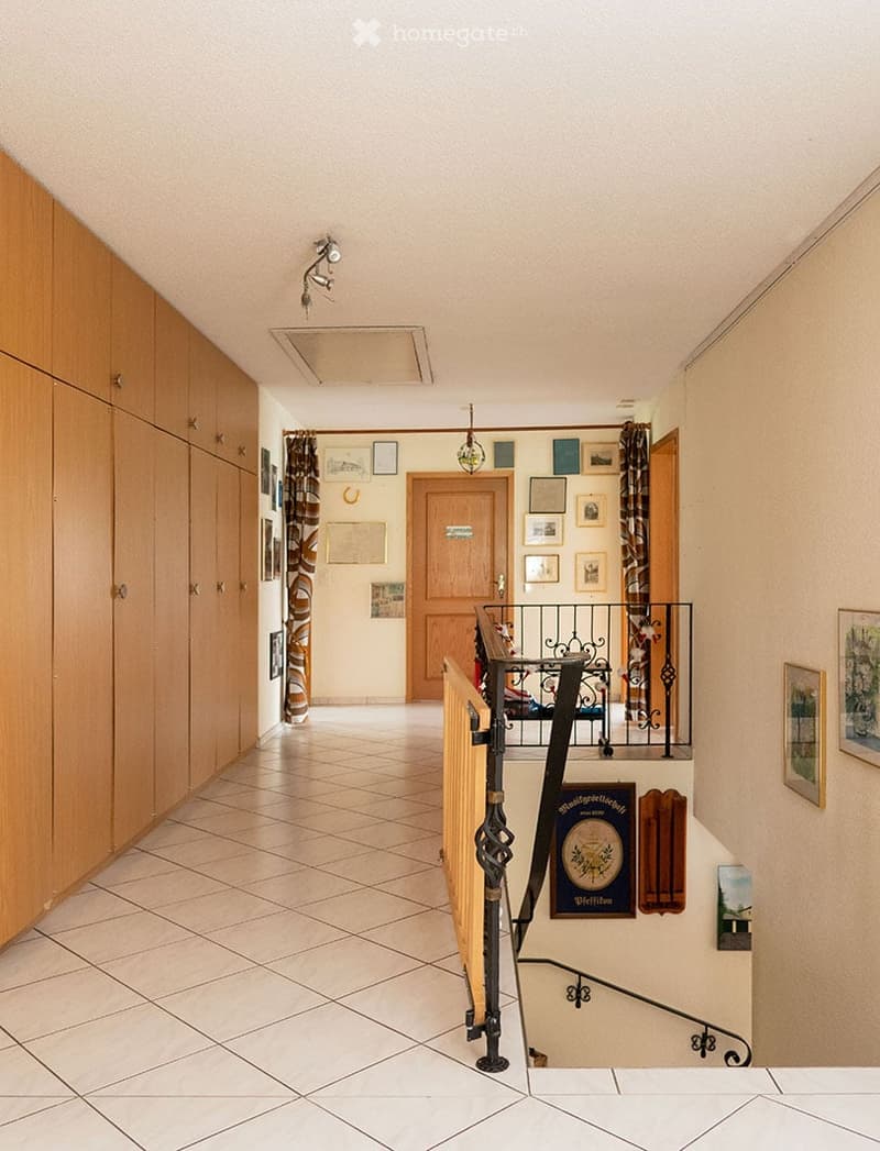 Spacious hallway with tiled flooring, wooden cabinetry on one side, and a staircase leading upwards. The walls are adorned with framed pictures and a decorative sign. The ceiling features recessed lighting and a central light fixture.
