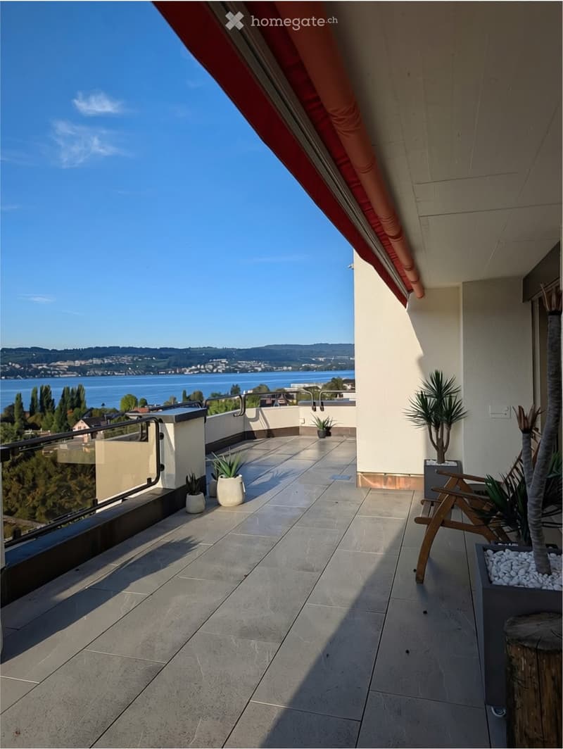 Balcony with tiled floor, wooden chair, potted plants, and scenic view of water and distant hills.