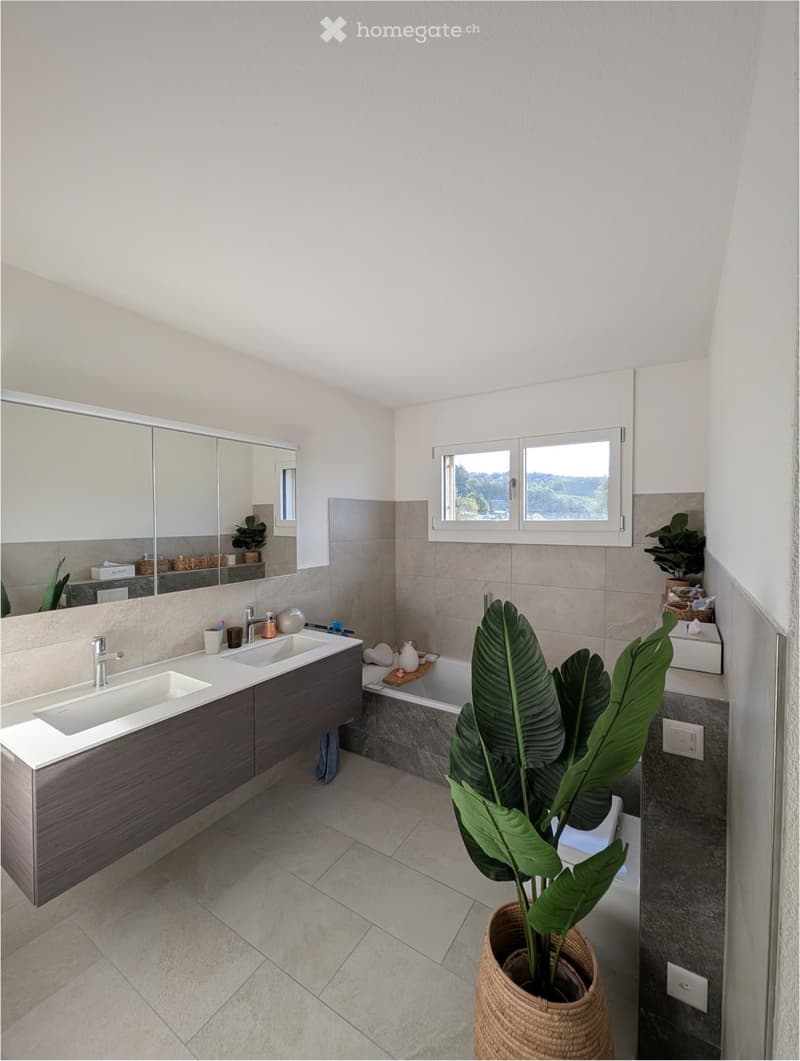 Modern bathroom with double sink, bathtub, large mirror, and potted plant near the window.