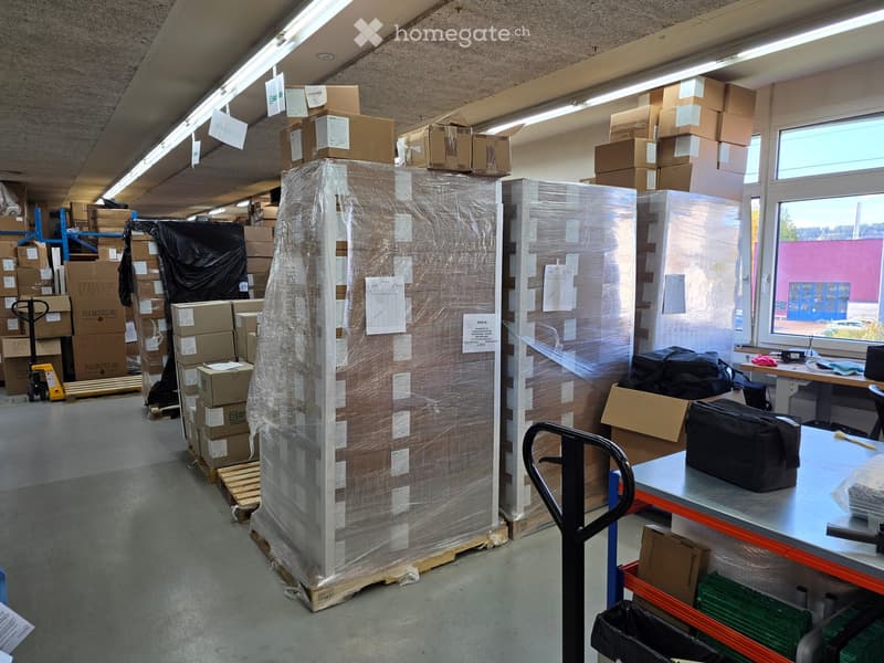 Warehouse space with stacked cardboard boxes, large wrapped pallets, a hand truck, and a table with a handbag on it.