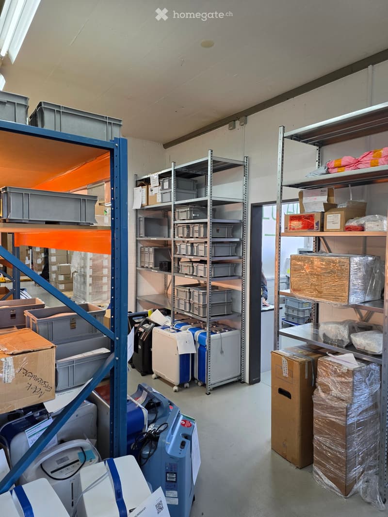 An empty storage room with metal shelving units, various boxes, and storage containers. The room has a high ceiling with exposed lighting fixtures and a concrete floor.