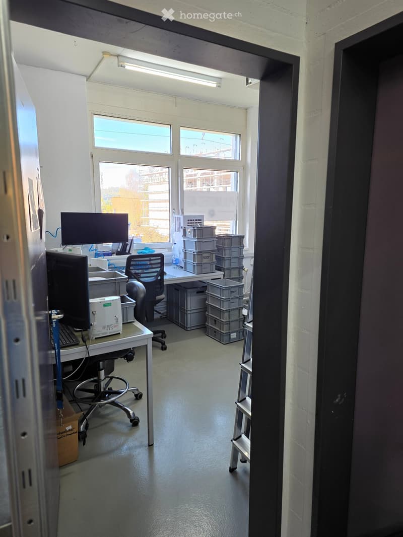 Office space with a desk, chair, screen, keyboard, and several stacked crates. The room has large windows and a door leading to another area.