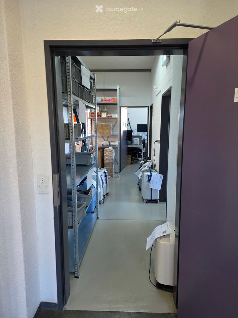 A clean, well-lit hallway with metal shelving units on the left side and various equipment and supplies on the right side, including a cart, a computer setup, and a white machine with papers attached.