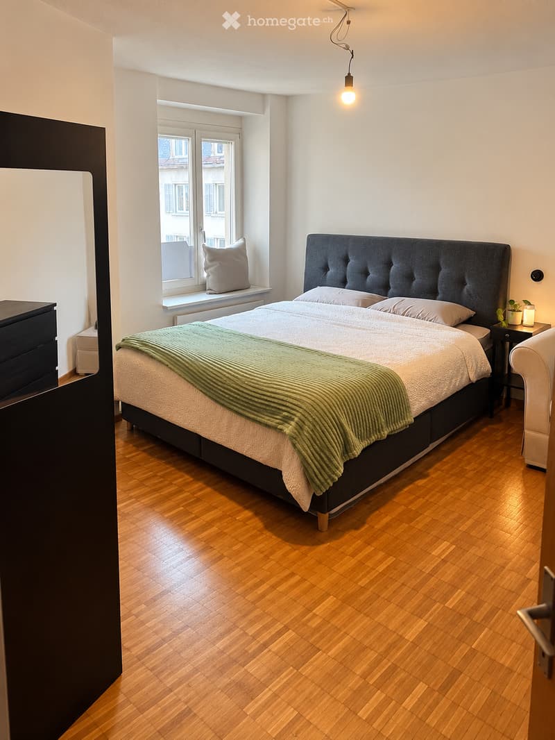 Modern bedroom with wooden flooring, a large bed with a tufted headboard, pillows, and a green blanket, a nightstand with a lamp and plant, and a mirror on the left side.