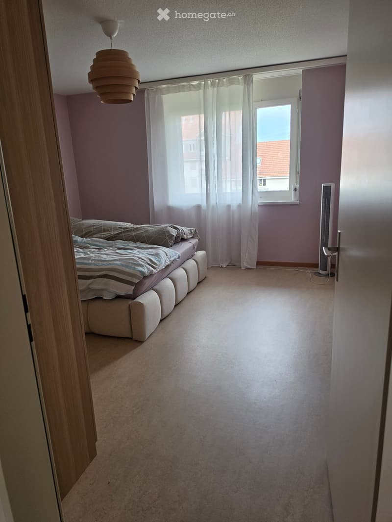 Bedroom with a bed, fan, and large window with curtains. Light purple walls and carpeted floor.