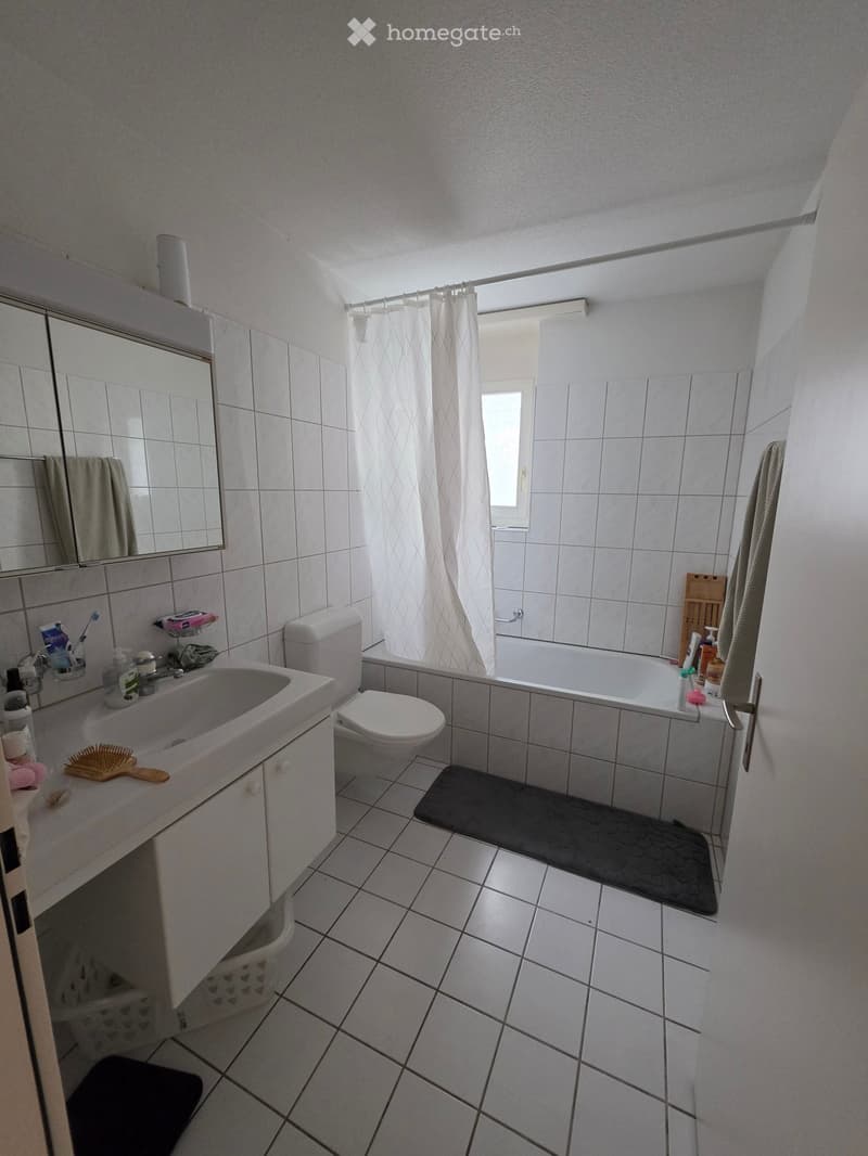 Bathroom with white tiles, a sink, toilet, bathtub, mirror, and a shower curtain. Includes a towel, bathroom mat, and personal care items on the sink counter.