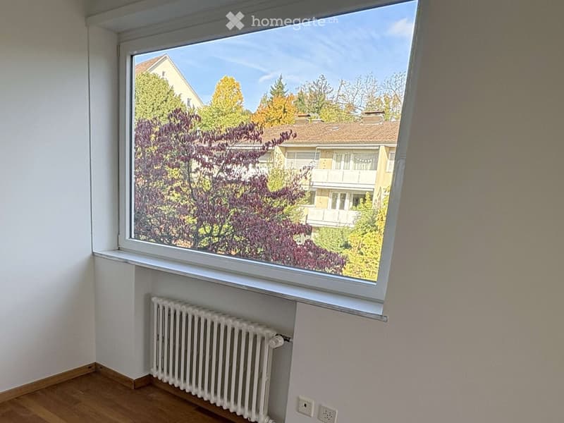 Empty room with wooden flooring, white walls, radiator, and large window showing a view of trees and neighboring houses.