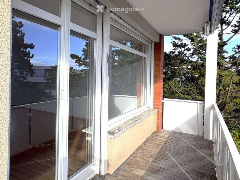 Balcony with wooden decking, white railing, large glass doors leading to the interior, and a view of greenery and trees.