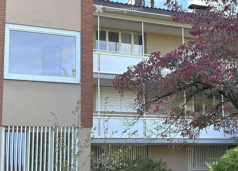 Multi-story residential building with balconies, large windows, and surrounding greenery.