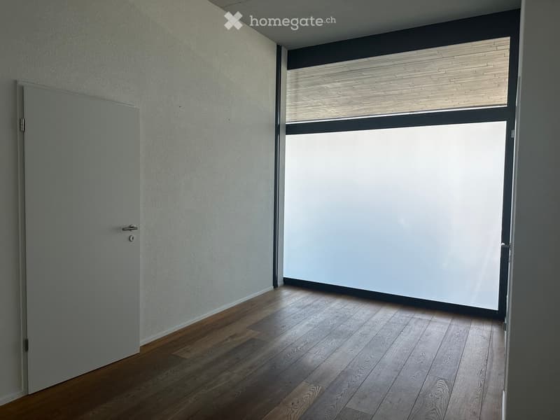 Empty room with wooden flooring, white walls, and a large window with blinds. The room features a closed door on the left side.