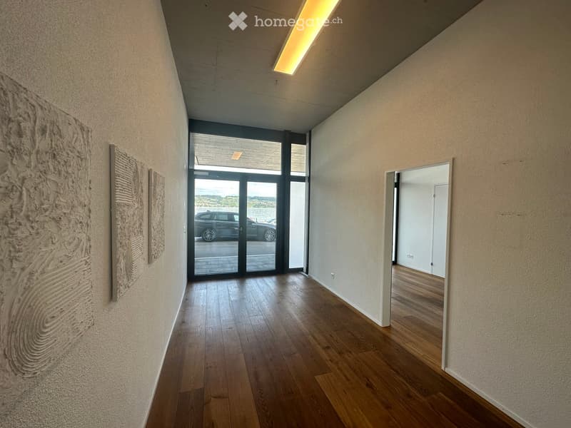 Empty hallway with wooden flooring, beige walls, and a large glass door leading outside. Artwork is displayed on one wall.