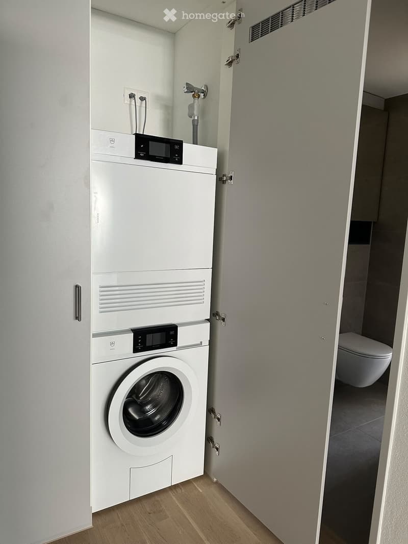 A compact laundry area with a stacked washing machine and dryer unit, a control panel on top, and an open door leading to a bathroom with a toilet visible.