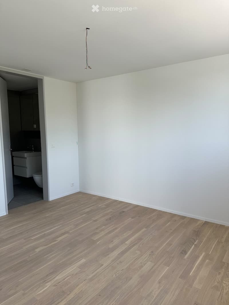 Empty room with light wooden flooring and white walls, featuring an open doorway leading to a bathroom with a sink and toilet.