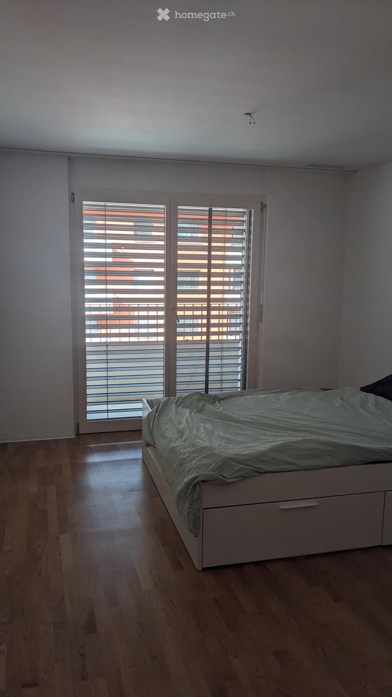 Minimalist bedroom with a single bed, light-colored bedding, and large windows with blinds. The floor is wooden, and the walls are white.