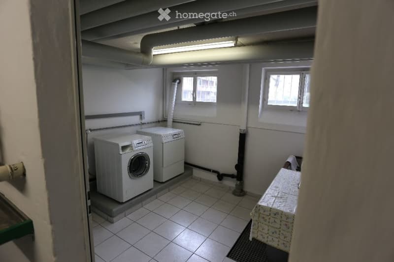 Utility room with white tiled floor, two white washing machines, and a small table with a patterned cloth.