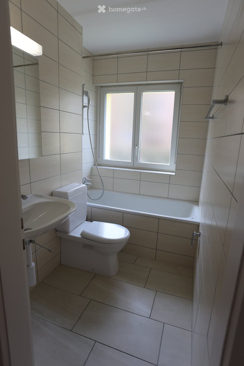 Modern bathroom with beige tiles, a sink, toilet, bathtub, and a window with frosted glass. The space is clean and well-lit.