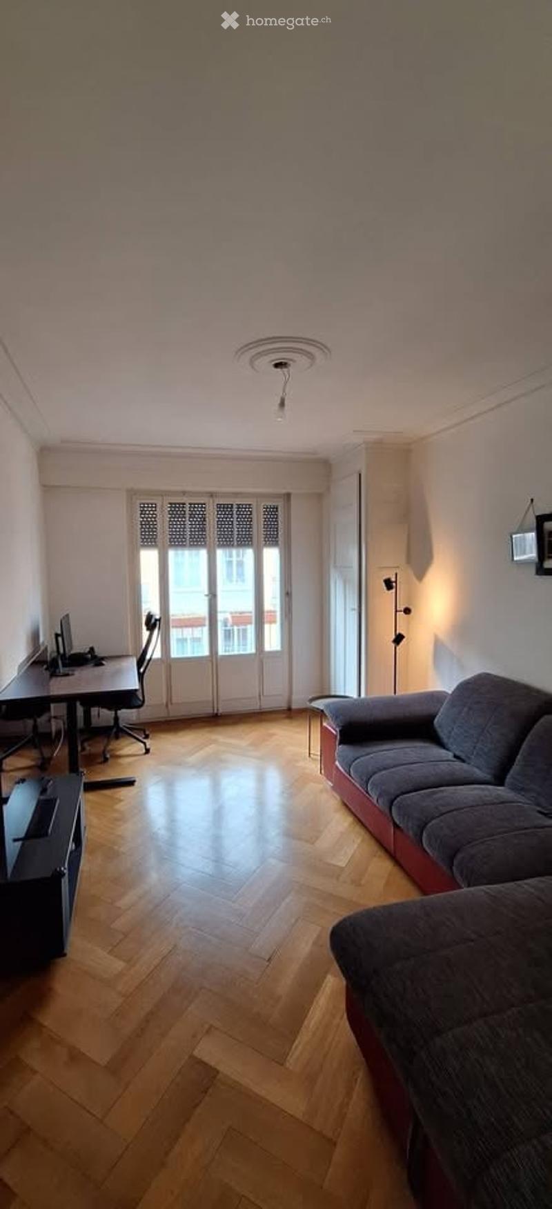 Bright, modern living room with wooden flooring, large window with shutters, grey sofa, black desk with computer, and a floor lamp.