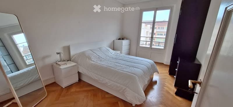 Bright bedroom with wooden flooring, large window offering a city view, a neatly made bed, a nightstand with a lamp, and a full-length mirror.