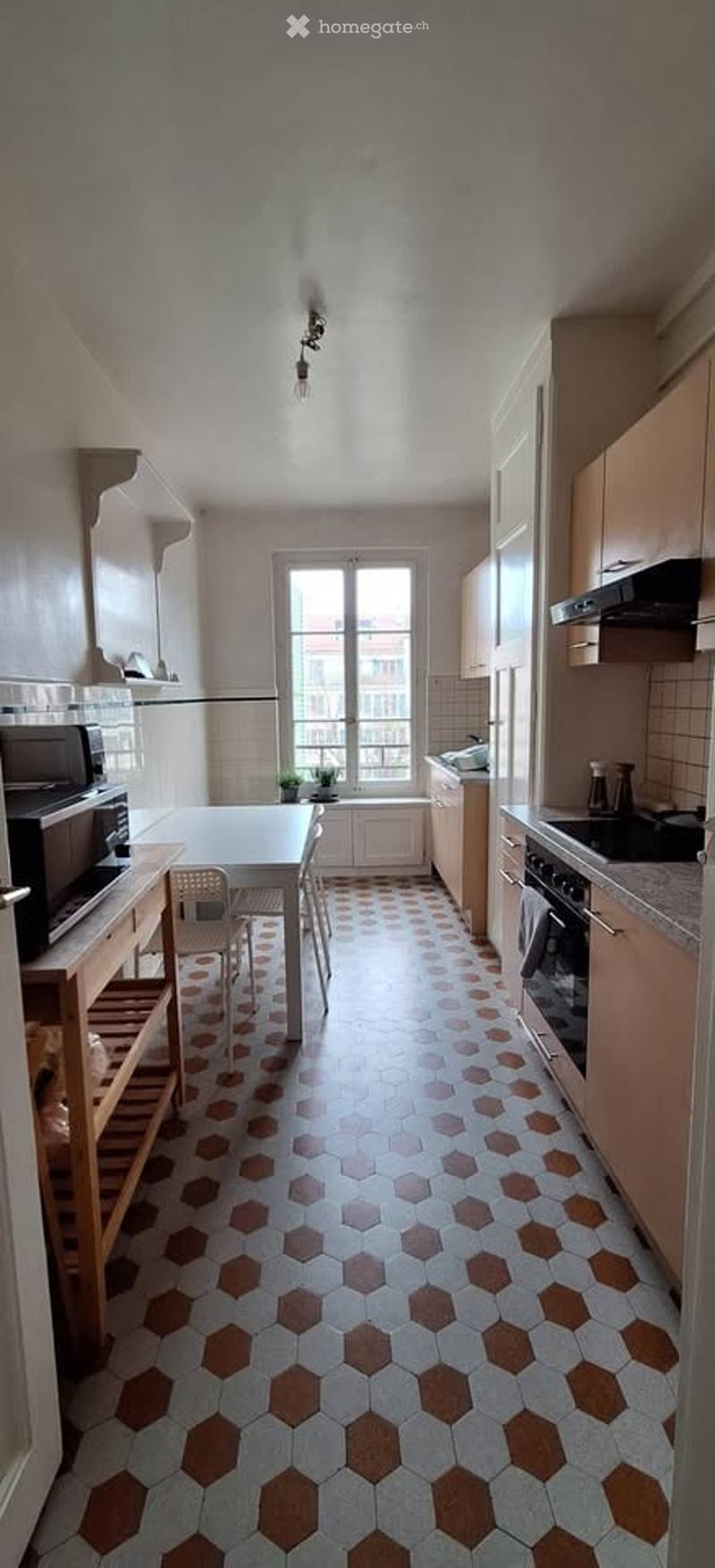 A well-lit kitchen with a hexagonal tile floor, wooden cabinets, and various appliances including a microwave, oven, and stove. A small dining area with a table and chairs is visible near the window.