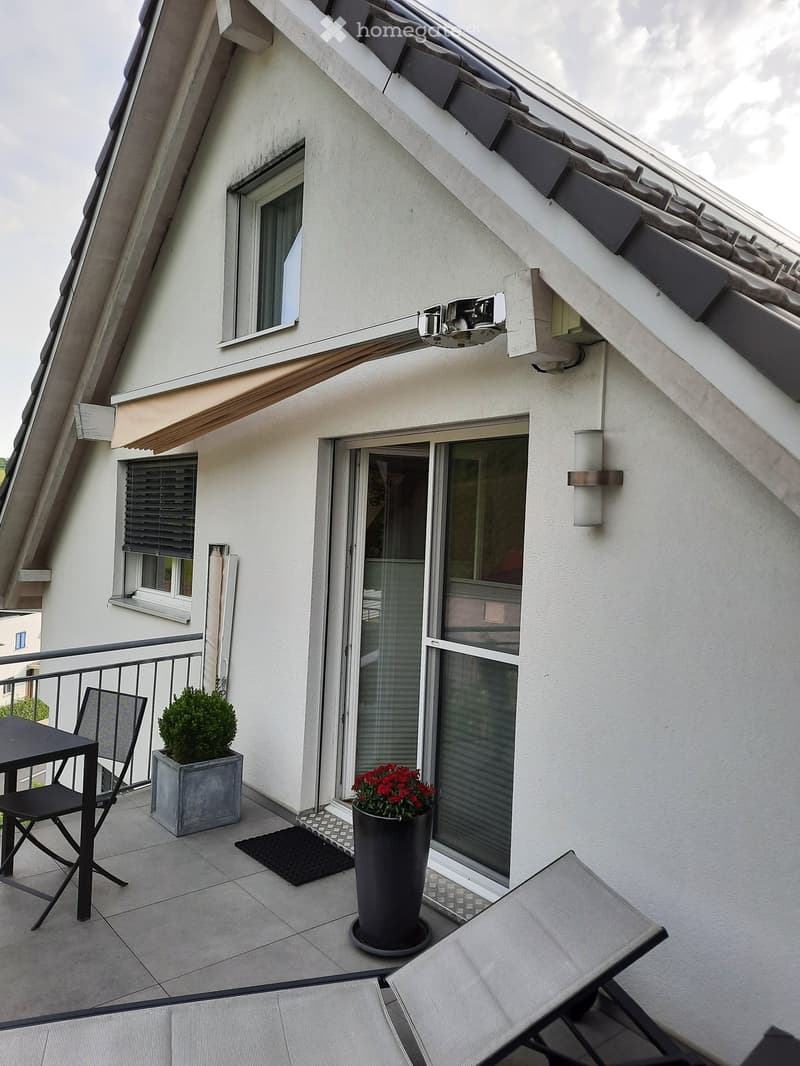 Balcony with tiled floor, black table and chair, potted plants, and a sliding glass door leading to the building.
