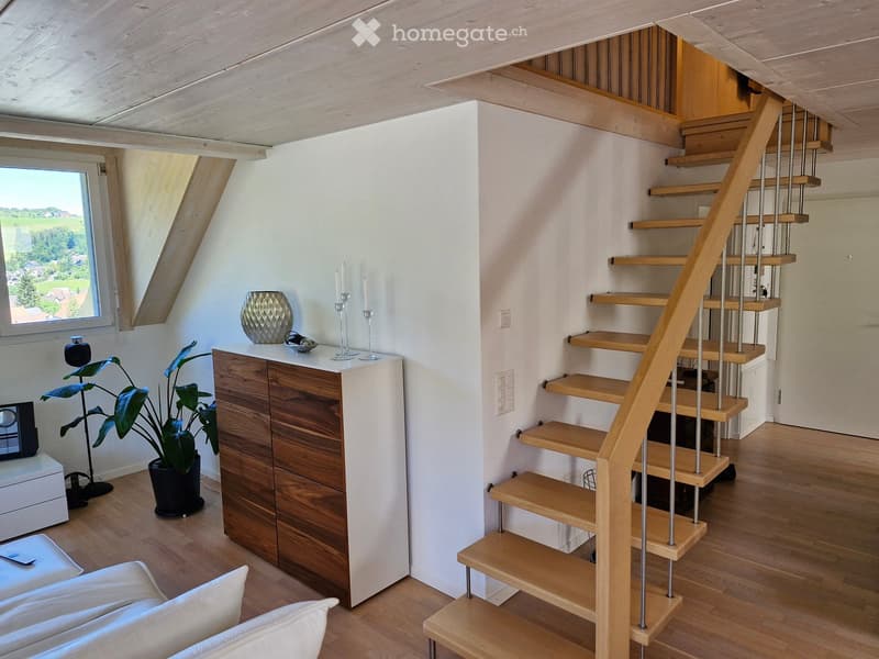 A modern, well-lit living space with a wooden staircase leading upwards, a white sofa, a wooden cabinet, a decorative vase, candle holders, a potted plant, and a speaker system.