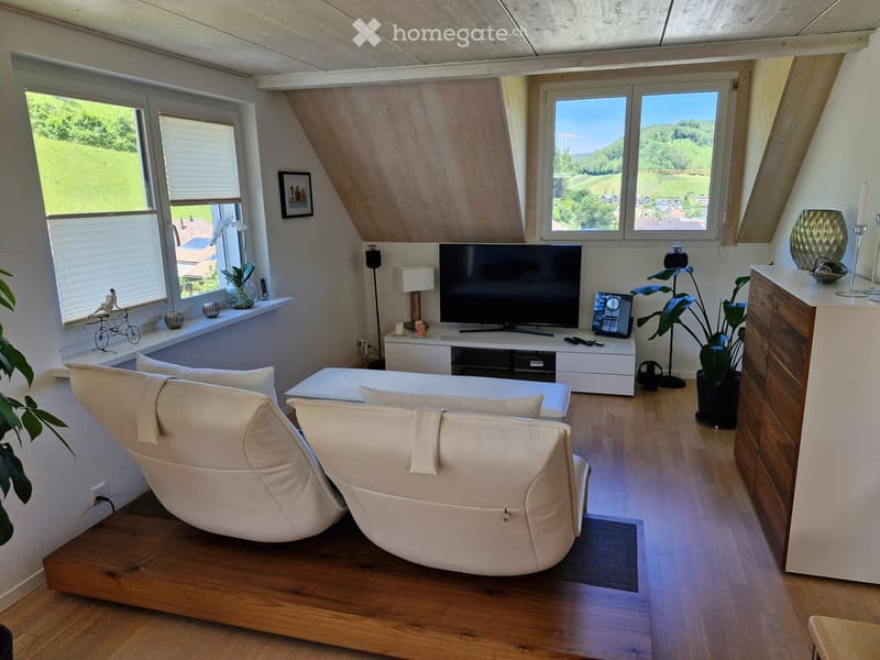Bright living room with wooden floors, large windows offering a scenic view, a white sectional sofa, a TV stand with a flat-screen TV, a side table with a lamp, and decorative plants.