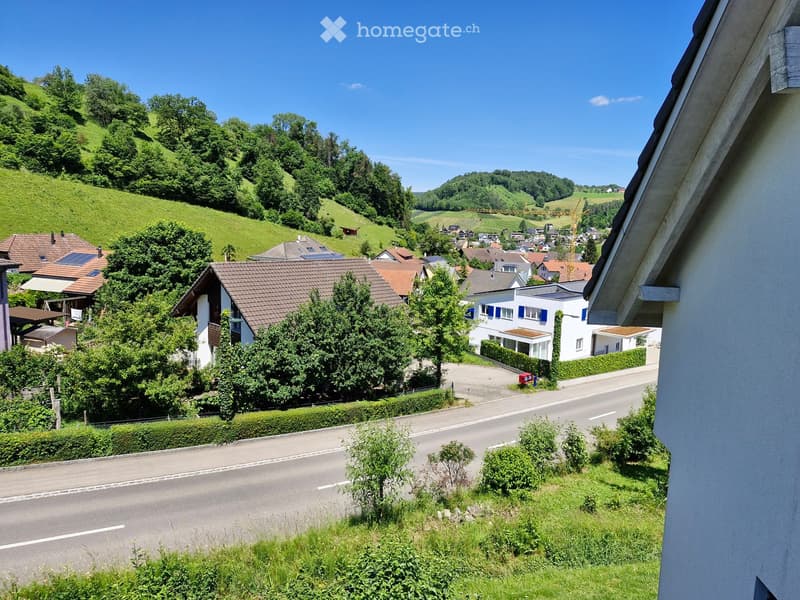 Residential area with houses, trees, and a road, surrounded by green hills and a clear blue sky.