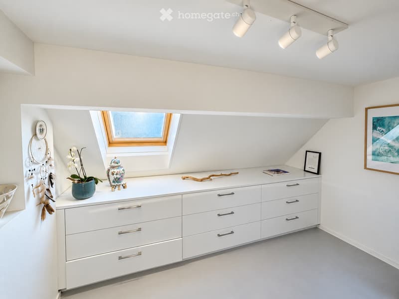 A modern, minimalist room with white walls, a large window, and a white cabinet with multiple drawers. The cabinet holds decorative items, including a vase with flowers, a dreamcatcher, and a framed picture.