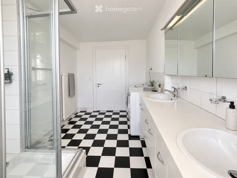 Modern bathroom with black and white checkered floor, white walls, large mirror, double sink, bathtub, and glass-enclosed shower. Includes towel, radiator, and small decorative items on the sink counter.