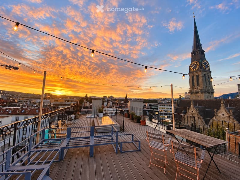 Rooftop terrace with wooden decking, string lights, a table with chairs, and a bench, overlooking a cityscape with a prominent clock tower at sunset.