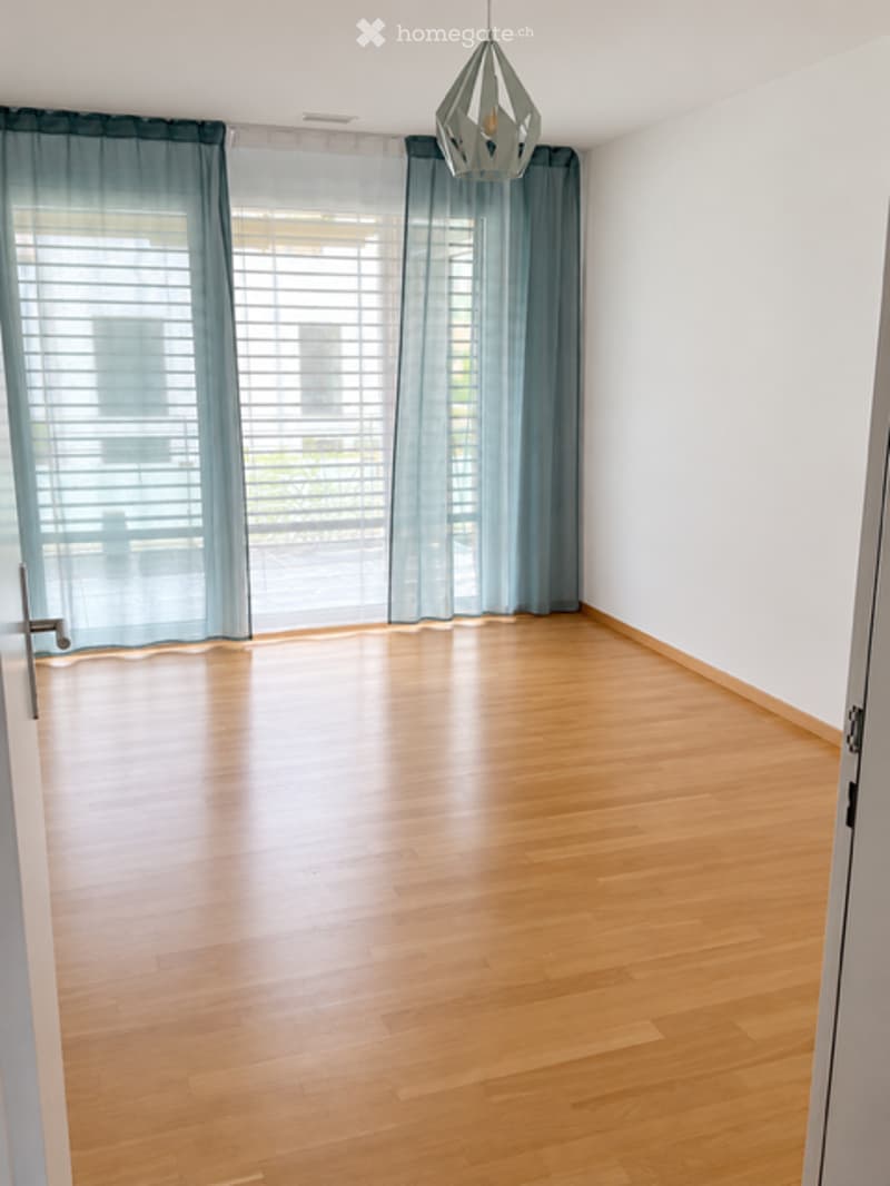 Empty room with wooden flooring, white walls, large windows with blue curtains, and a modern hanging light fixture.