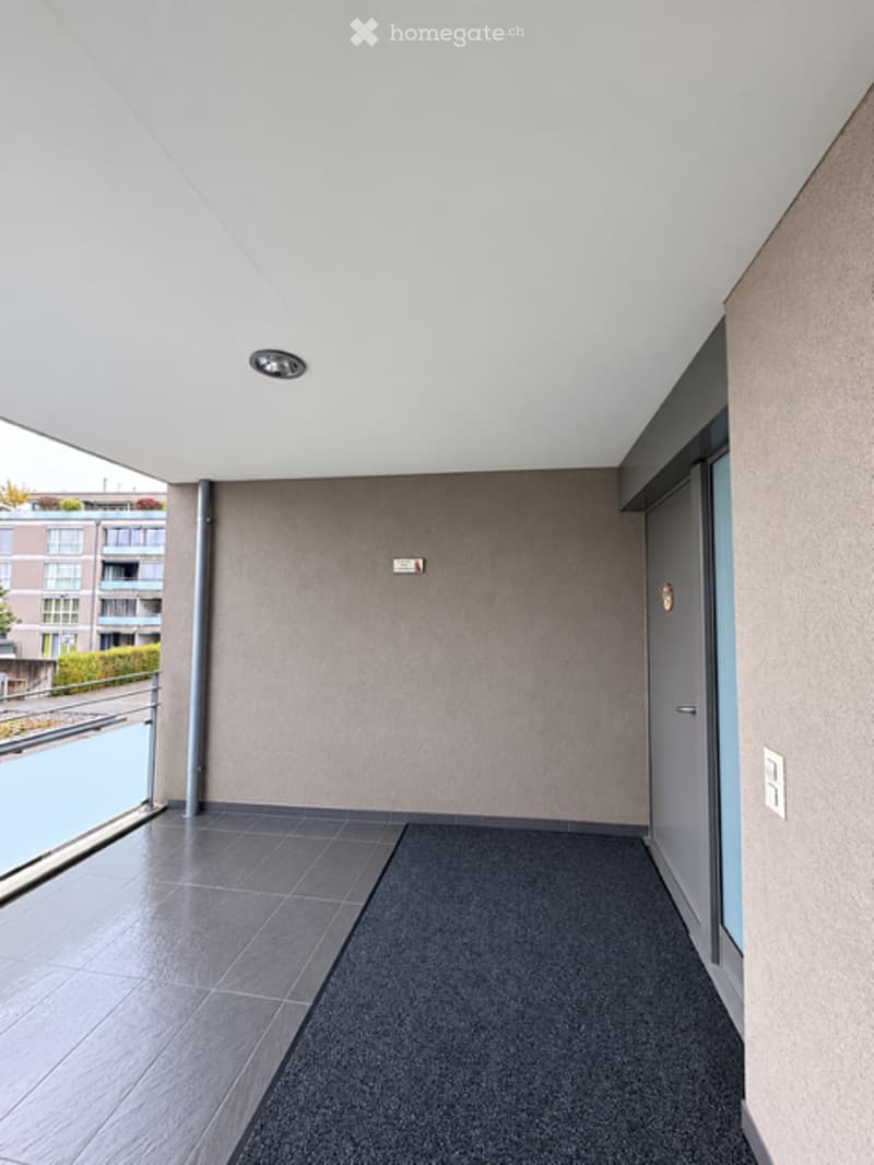 Modern balcony with tiled floor, grey walls, and a glass railing overlooking a cityscape.