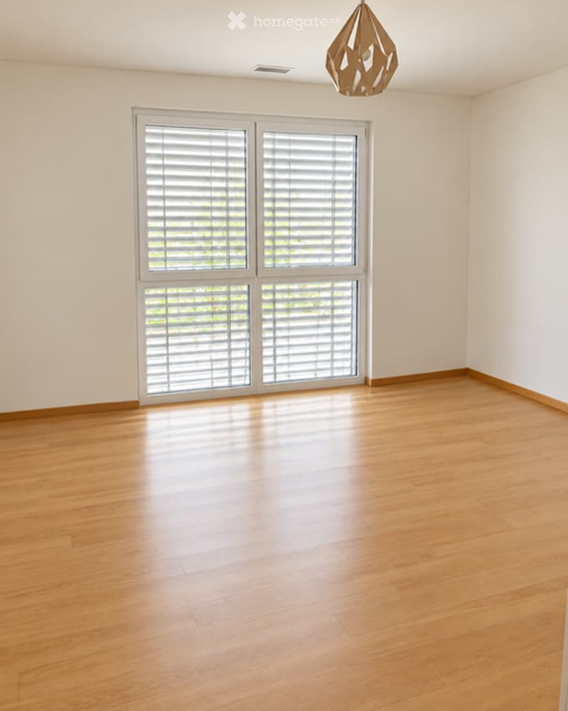 Empty room with wooden flooring, large window with blinds, and a hanging light fixture.