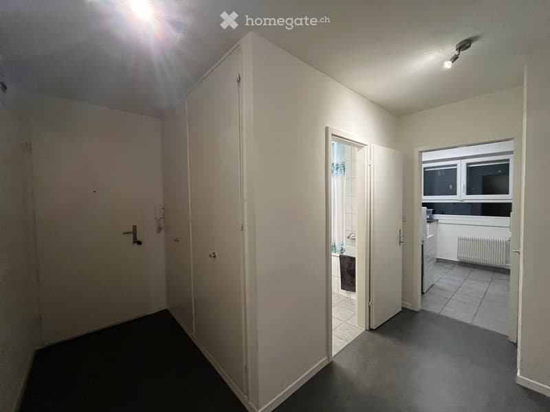 Empty hallway with white walls, dark flooring, and two doors leading to a bathroom and kitchen area.