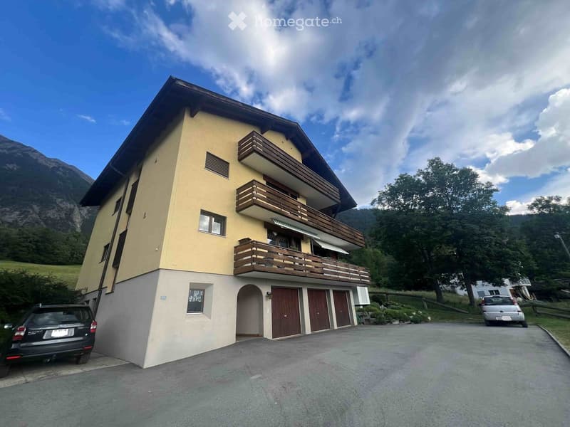 Two-story house with beige walls, wooden balconies, and a garage. Located in a scenic area with mountains and trees in the background.