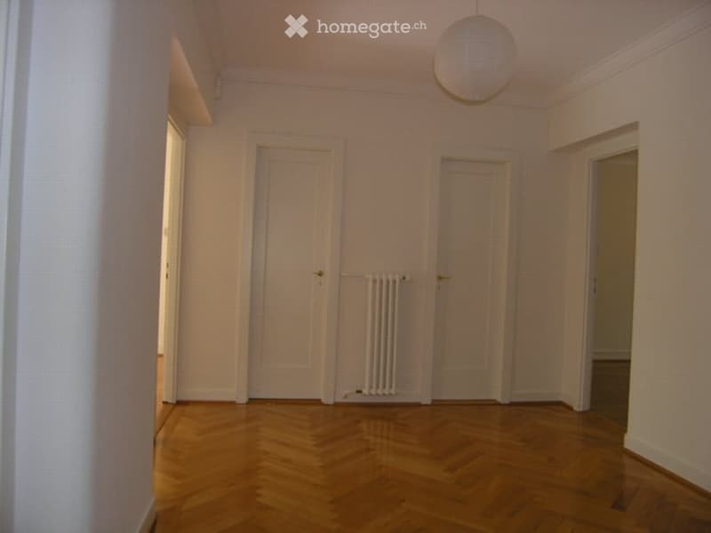Empty room with wooden flooring, white walls, and three doors. A radiator is visible under one door, and a hanging light fixture is on the ceiling.