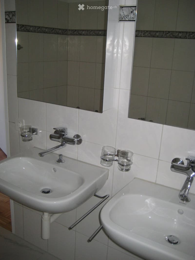 Modern bathroom with two white sinks, chrome faucets, mirrors above, and two glass cups mounted on the wall.