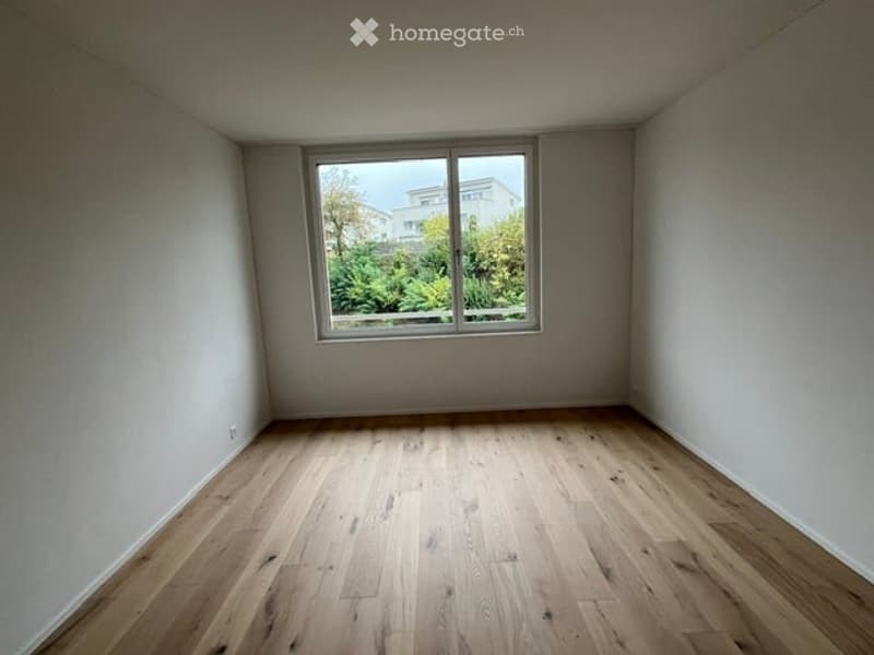 Empty room with wooden flooring and a large window showing greenery outside.