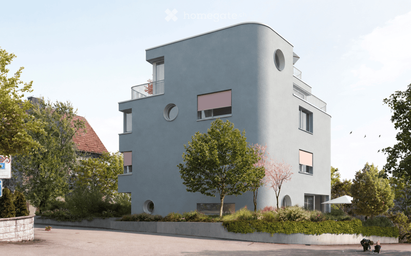 Modern, two-story building with a sleek, curved facade in light blue, featuring large windows and balconies. The building is surrounded by greenery and trees, with a raised ground floor and a small garden area in front.