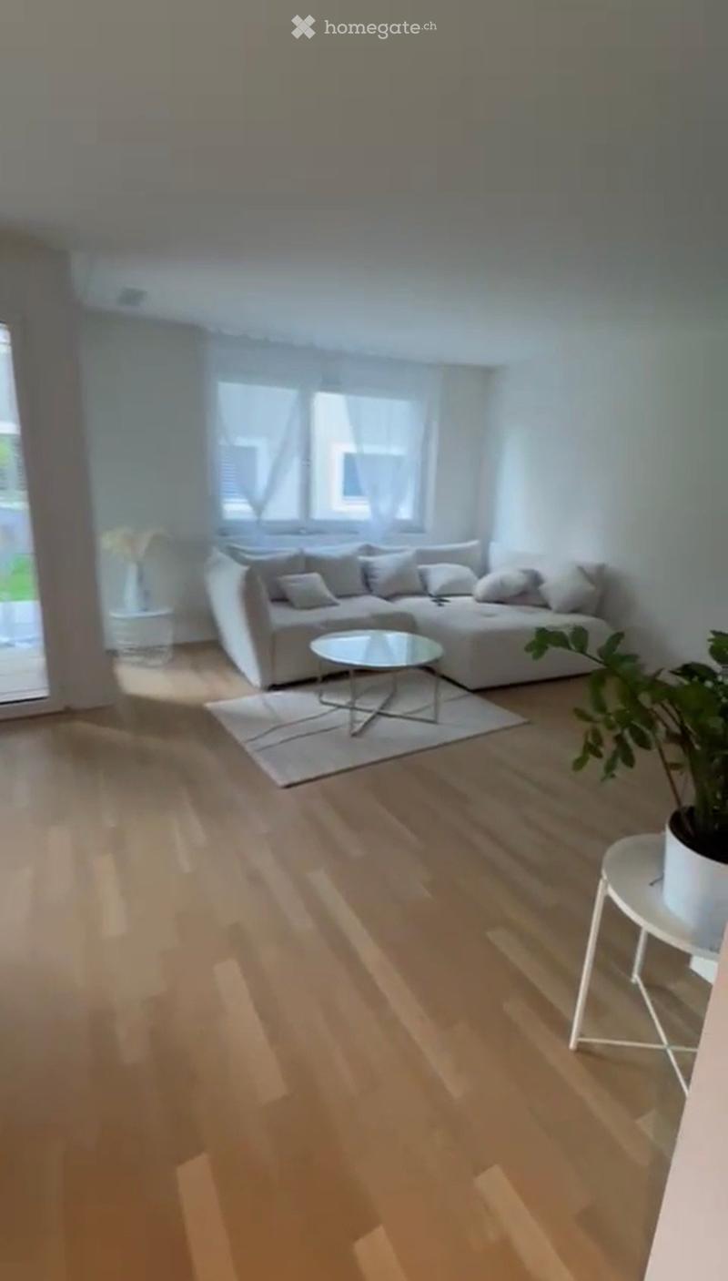 Modern living room with light wooden flooring, a grey sectional sofa, a round glass coffee table, a side table with a plant, and large windows with sheer curtains.