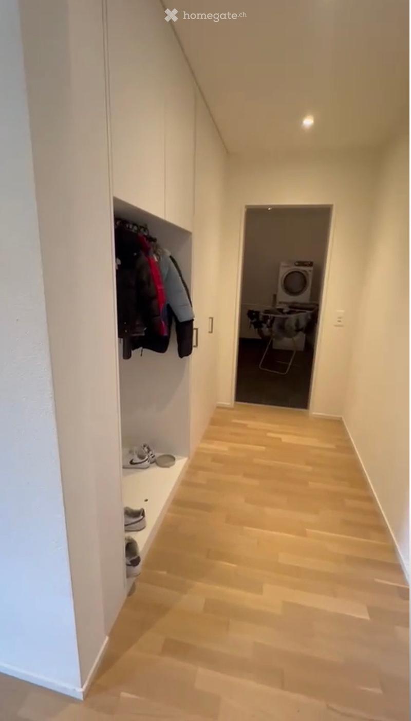 A narrow hallway with light wooden flooring, white walls, and built-in storage shelves holding shoes and jackets. A doorway leads to a laundry area with a washing machine and dryer visible.