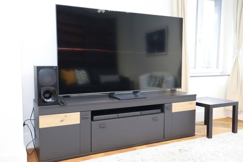 Modern living room with a large flat-screen TV on a sleek entertainment unit, accompanied by a speaker and a small side table.