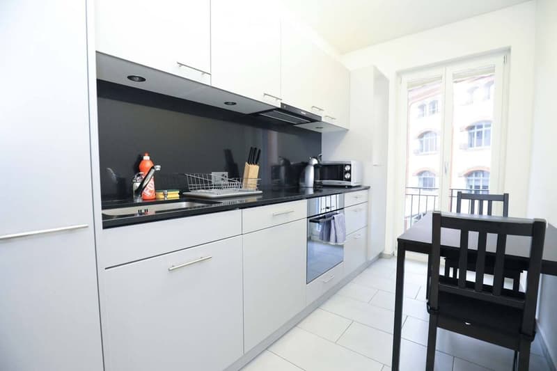 Modern kitchen with white cabinets, black countertops, and stainless steel appliances. Includes a sink, faucet, dish rack, oven, microwave, and a small dining area with a table and chairs.