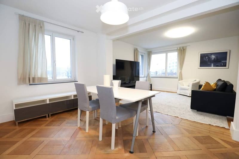 A modern, well-lit living and dining area with wooden flooring, large windows, a white dining table with four chairs, a black sofa with cushions, a television, and a picture frame on the wall.