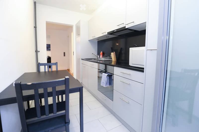 Modern kitchen with white cabinets, black countertops, and stainless steel appliances. Includes a sink, microwave, oven, and a small dining area with a table and chairs.