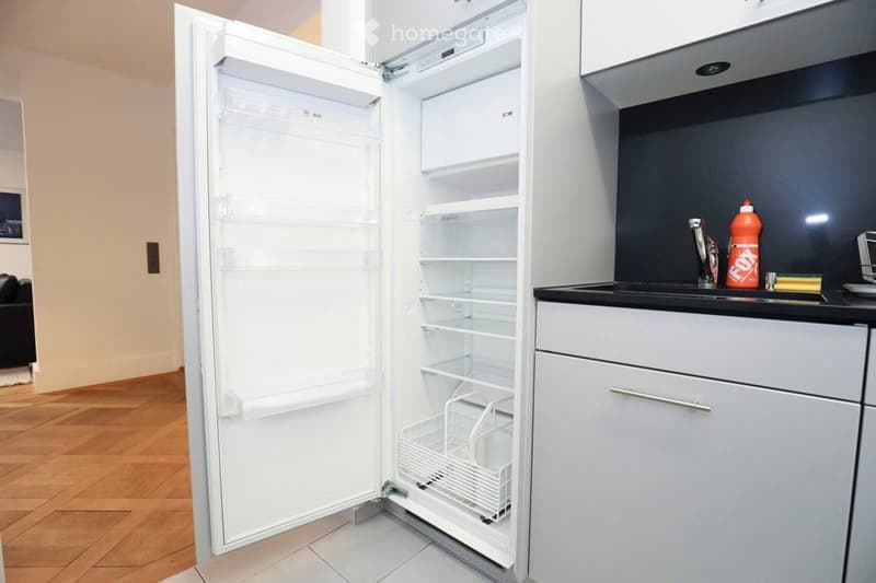 Modern kitchen with white cabinets, a stainless steel sink, and an open refrigerator. The floor is wooden, and the countertops are black.